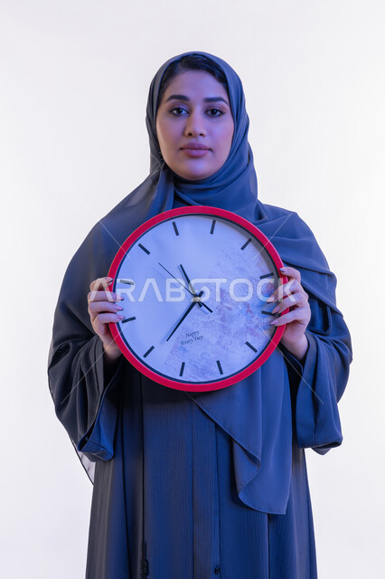 إنجاز المهام في الموعد المحدد, التذكيرات و التنبيهات الثابتة, بورتريه مقرب لامرأة عربية خليجية إماراتية ترتدي العباءة تمسك بيدها ساعة حائط حمراء, شابة سعودية تنظر الى الكاميرا بإيماءات الفخر والسرور، أهمية الحفاظ على الأوقات، خلفية بيضاء