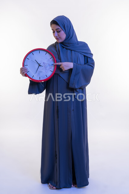 إنجاز المهام في الموعد المحدد, التذكيرات و التنبيهات الثابتة, أهمية الحفاظ على الأوقات, بورتريه لامرأة عربية خليجية إماراتية ترتدي العباءة تمسك بيدها ساعة حائط حمراء، شابة سعودية تشير بإصبع السبابة إلى وقت معين, الجسم بالطول الكامل, خلفية بيضاء