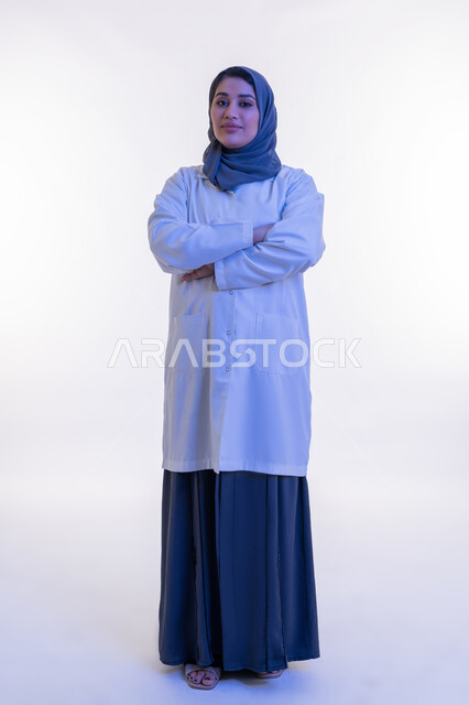 النظر إلى الكاميرا بتعابير الثقة بالنفس والسرور، تقديم الخدمات العلاجية والفحوصات الطبية للمرضى, بورتريه  لطبيبة عربية خليجية اماراتية محجبة ترتدي المعطف الطبي الأبيض تقف بإيماءات أيدي متقاطعة, شابة سعودية تعمل في القطاع الصحي، الجسم الكامل بالطول، خلفية بيضاء