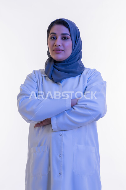 العمل في القطاع الصحي، تقديم الخدمات العلاجية والفحوصات الطبية للمرضى, بورتريه مقرب  لطبيبة عربية خليجية اماراتية محجبة ترتدي المعطف الطبي الأبيض تقف بإيماءات أيدي متقاطعة، شابة سعودية تنظر إلى الكاميرا بتعابير الثقة بالنفس والسرور، خلفية بيضاء