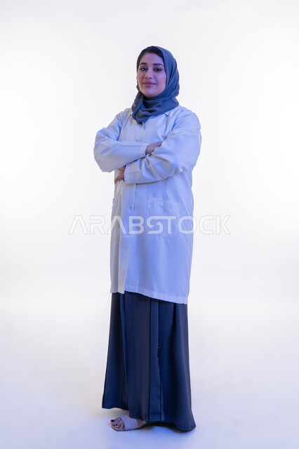 النظر إلى الكاميرا بتعابير الثقة بالنفس والسرور، تقديم الخدمات العلاجية والفحوصات الطبية للمرضى, بورتريه  لطبيبة عربية خليجية اماراتية محجبة ترتدي المعطف الطبي الأبيض تقف بإيماءات أيدي متقاطعة, شابة سعودية تعمل في القطاع الصحي، الجسم الكامل بالطول، خلفية بيضاء
