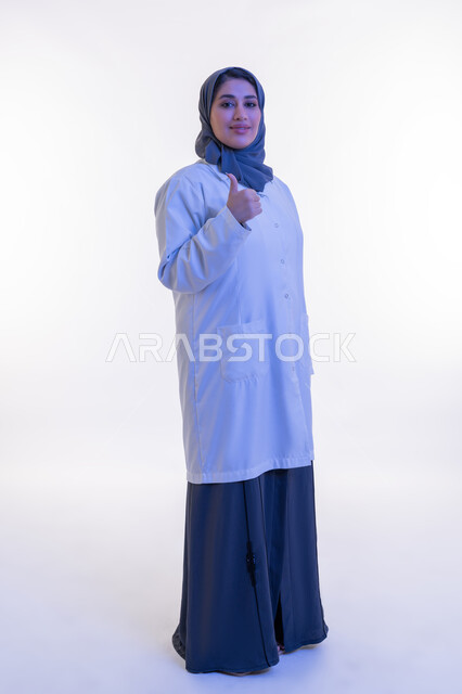 تقديم الخدمات العلاجية والفحوصات الطبية للمرضى، شابة سعودية تعمل في القطاع الصحي, بورتريه لطبيبة عربية خليجية اماراتية محجبة مبتسمة ترتدي المعطف الطبي الأبيض ترفع اصبع الابهام بتعابير الاعجاب والموافقة, الجسم الكامل بالطول، خلفية بيضاء