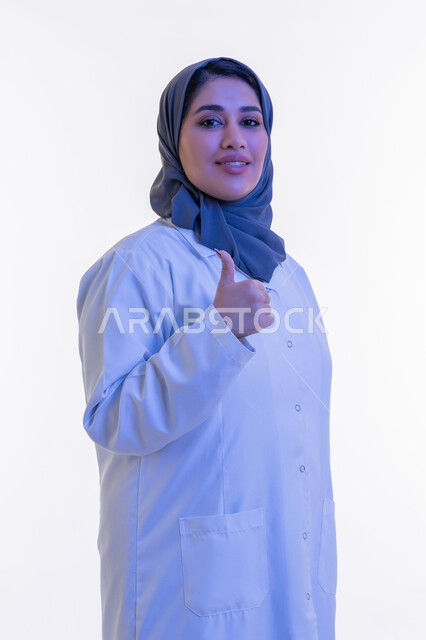 تقديم الخدمات العلاجية والفحوصات الطبية للمرضى، شابة سعودية تعمل في القطاع الصحي, بورتريه مقرب لطبيبة عربية خليجية اماراتية محجبة مبتسمة ترتدي المعطف الطبي الأبيض ترفع اصبع الابهام للاعلى بتعابير الاعجاب والموافقة، خلفية بيضاء
