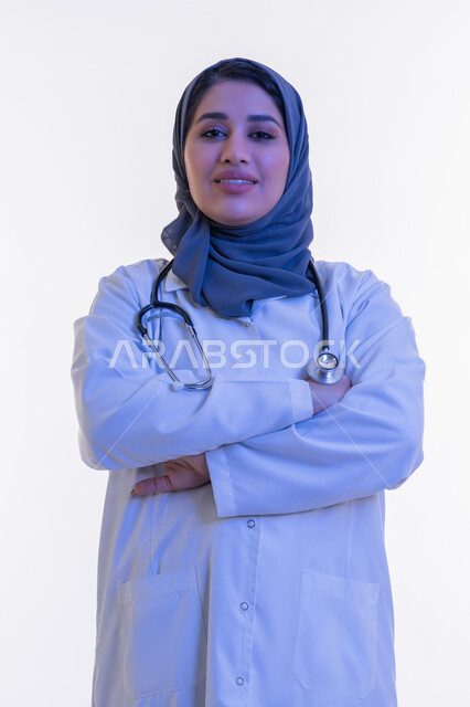 Providing medical services and examinations to patients, working in the health sector, close-up portrait of an Arab Gulf Emirati female doctor wearing a white medical coat and an examination stethoscope standing with crossed hands gestures, a Saudi woman looking at the camera with an expression of self-confidence, white background