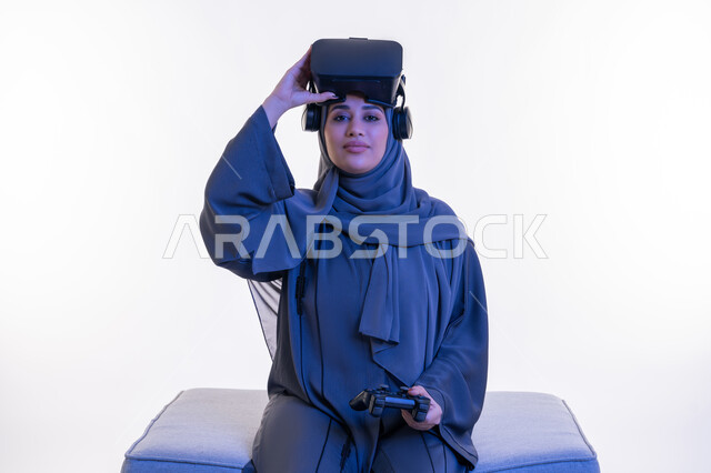 Playing video games, a young Saudi woman raising her glasses to the imaginary world and looking at the camera with an expression of pleasure, using modern devices, a portrait of an Arab Gulf Emirati woman wearing an abaya and virtual reality glasses sitting on a comfortable sofa holding a joystick in her hand, white background