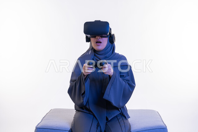 استخدام الأجهزة الحديثة، دمج التسلية مع العالم الخيالي، بورتريه لإمرأة عربية خليجية إماراتية ترتدي العباءة ونظارة الواقع الإفتراضي تجلس على أريكة مريحة وتمسك بيدها عصا التحكم بتعابير الاندهاش والذهول، شابة سعودية تلعب بألعاب الفيديو الترفيهية، خلفية بيضاء