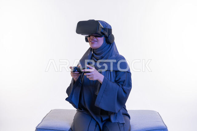Using modern devices and technologies, combining entertainment with the imaginary world, portrait of an Arab Gulf Emirati woman wearing an abaya and virtual reality glasses sitting on a comfortable sofa and holding a joystick in her hand with happy expressions, a young Saudi woman playing recreational video games, white background