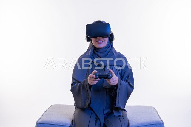 Expressions of happiness and pleasure, combining entertainment with the imaginary world, portrait of a veiled Gulf Arab Emirati woman wearing an abaya and virtual reality glasses sitting on a comfortable sofa and holding a joystick in her hand, a young Saudi woman playing recreational video games, white background
