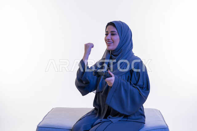 مفهوم استخدام التقنية الحديثة، قضاء اوقات ممتعة  بألعاب الفيديوز الترفيهية في عطلة نهاية الاسبوع، بورتريه لإمرأة عربية خليجية إماراتية ترتدي العباءة تجلس على اريكة مريحة تحمل بيدها عصا التحكم، شابة سعودية تغلق قبضة يدها بتعابير الفوز والنصر والقوة، خلفية بيضاء