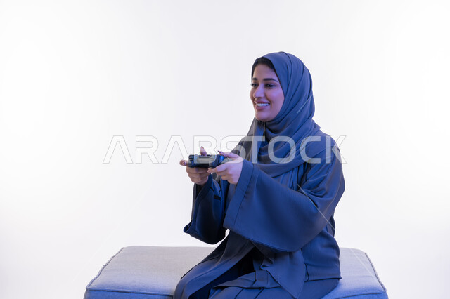 ايماءات الاندماج والتفاعل، شابة سعودية تلعب بألعاب الفيديوز الترفيهية في عطلة نهاية الاسبوع، استخدام التقنيات الالكترونية الحديثة والمتطورة، بورتريه لإمرأة عربية خليجية إماراتية مبتسمة ترتدي العباءة تجلس على اريكة مريحة تحمل بيدها عصا التحكم بتعابير السعادة، خلفية بيضاء