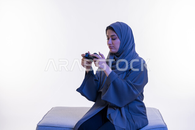 Using modern and advanced electronic technologies, a young Saudi woman playing recreational video games on the weekend, a side portrait of an Arab Gulf Emirati woman wearing an abaya sitting on a comfortable sofa holding a joystick in her hand and looking at it with thinking gestures, white background