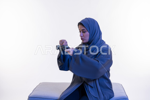 قضاء اوقات ممتعة  بألعاب الفيديوز الترفيهية في عطلة نهاية الاسبوع، استخدام التقنيات الالكترونية الحديثة والمتطورة، بورتريه من الجانب لإمرأة عربية خليجية إماراتية ترتدي العباءة تجلس على اريكة مريحة تحمل بيدها عصا التحكم، شابة سعودية تنظر للقبضة، خلفية بيضاء