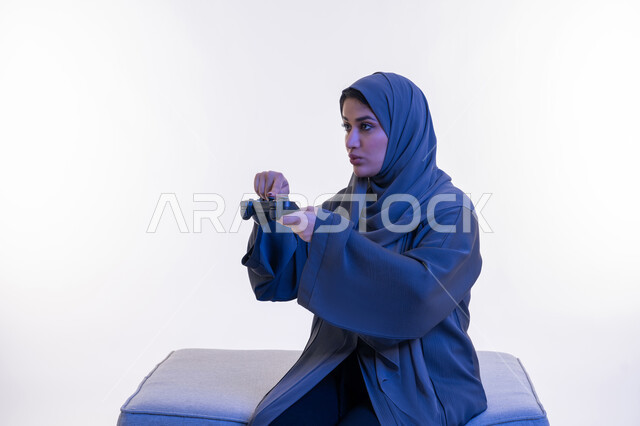 ايماءات الاندماج والتفاعل، قضاء اوقات ممتعة  بألعاب الفيديوز الترفيهية في عطلة نهاية الاسبوع، شابة سعودية تستخدم التقنية الحديثة، بورتريه لإمرأة عربية خليجية إماراتية ترتدي العباءة  تجلس على اريكة مريحة تحمل بيدها عصا التحكم بتعابير السعادة، خلفية بيضاء