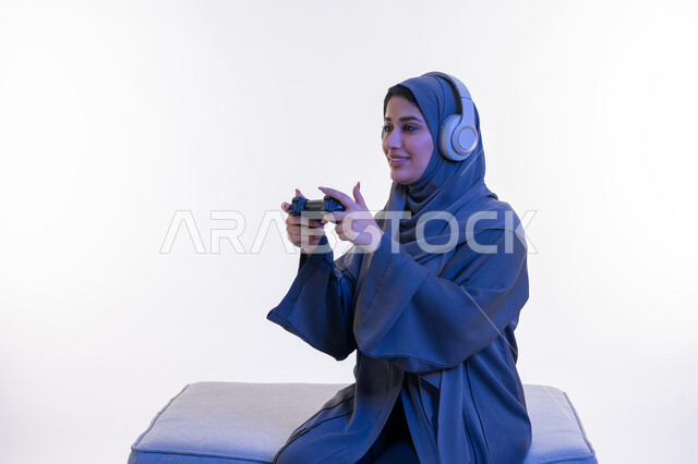 Gestures of integration and interaction during play, a young Saudi woman playing recreational video games, using modern and advanced electronic technologies, a side portrait of an Arab Gulf Emirati woman wearing an abaya and headphones sitting on a comfortable sofa holding a joystick in her hand, white background
