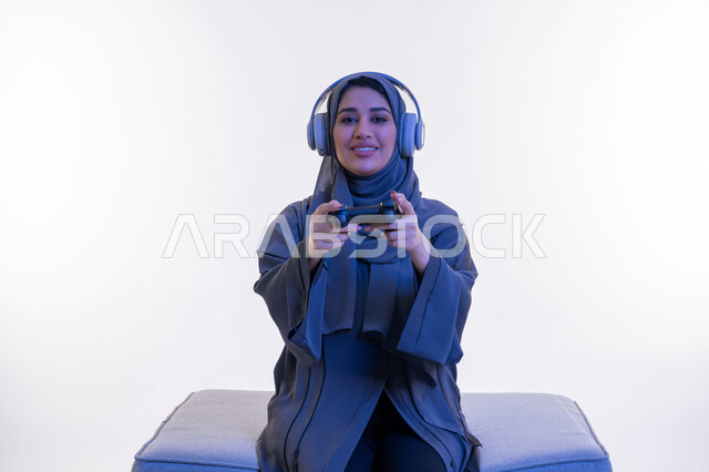 Gestures of integration and interaction during play, a young Saudi woman playing recreational video games on the weekend, using modern and advanced electronic technologies, a portrait of an Arab Gulf Emirati woman wearing an abaya and a headset holding a joystick in her hand, white background