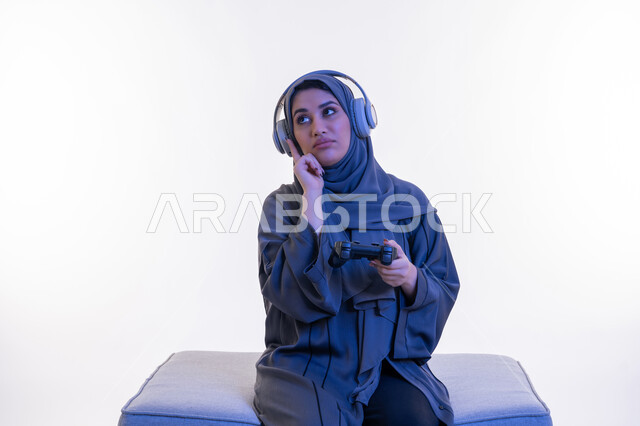 Using modern and advanced electronic technology, portrait of an Arab Gulf Emirati woman wearing an abaya and headphones sitting on a comfortable sofa holding a joystick in her hand, a young Saudi woman putting her hand on her cheek with a thinking expression, spending fun times playing entertaining video games, white background