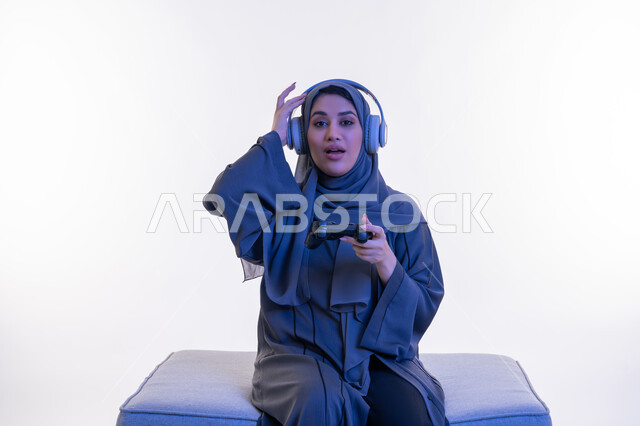 قضاء اوقات ممتعة  بألعاب الفيديوز الترفيهية، استخدام التقنية الحديثة والمتطورة، بورتريه  لإمرأة عربية خليجية إماراتية ترتدي العباءة وسماعة الرأس تجلس على اريكة مريحة تحمل بيدها عصا التحكم، شابة سعودية تضع يدها على رأسها بتعابير الخسارة والاندهاش، خلفية بيضاء