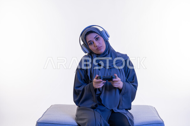 ايماءات الاندماج والتفاعل أثناء اللعب، شابة سعودية تلعب بألعاب الفيديوز الترفيهية، استخدام التقنيات الالكترونية الحديثة والمتطورة، بورتريه  لإمرأة عربية خليجية إماراتية ترتدي العباءة وسماعة الرأس تجلس على اريكة مريحة تحمل بيدها عصا التحكم، خلفية بيضاء