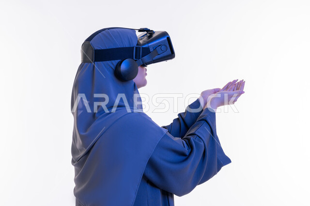 دمج التسلية بالتقنية الحديثة، شابة سعودية ترفع يديها لمحاولة حمل مجسم شيء ما، المتعة بالمشاهدة عبر نظارة الواقع الافتراضي, بورتريه مقرب من الجانب لإمرأة عربية خليجية إماراتية محجبة ترتدي العباءة تضع نظارات الفي آر, الاندماج ومحاكاة العالم الخيالي، خلفية بيضاء