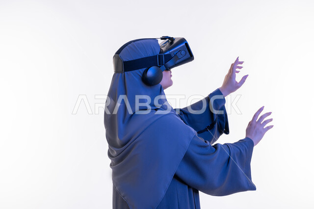 الاندماج ومحاكاة العالم الخيالي، المتعة و التسلية بالمشاهدة عبر نظارة الواقع الافتراضي، بورتريه مقرب من الجانب لإمرأة عربية خليجية إماراتية محجبة ترتدي العباءة وتنظر من خلال نظارات ثلاثية الأبعاد، شابة سعودية ترفع كف يديها للاعلى لمحاولة لمس شيء ما، خلفيه بيضاء