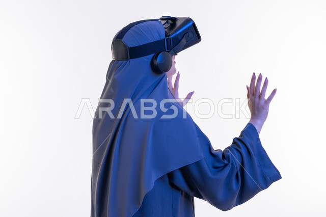 محاكاة العالم الخيالي، التسلية بالمشاهدة عبر نظارة الواقع الافتراضي، بورتريه مقرب من الجانب لإمرأة عربية خليجية إماراتية محجبة ترتدي العباءة تنظر من خلال نظارات ثلاثية الأبعاد، شابة سعودية ترفع كف يديها للاعلى لمحاولة لمس شيء ما بتعابير الاندماج، خلفيه بيضاء