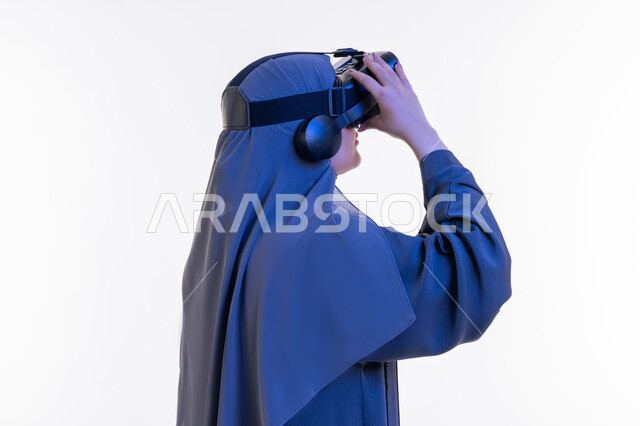 Fun and entertainment watching through virtual reality glasses, close-up portrait from the side of a veiled Arab Gulf Emirati woman wearing an abaya and looking through 3D glasses, a young Saudi woman using modern technology, merging and simulating the imaginary world, white background