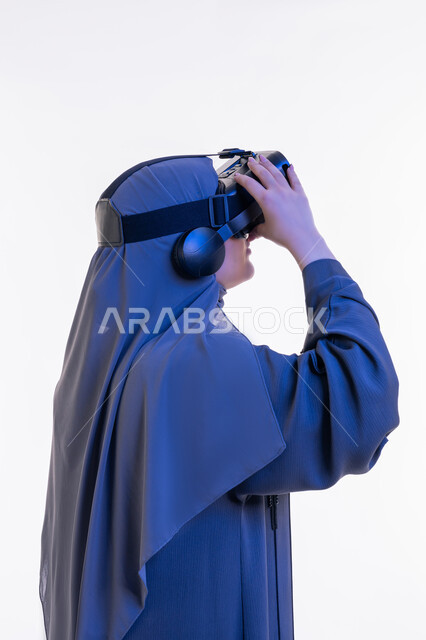 Fun and entertainment watching through virtual reality glasses, close-up portrait from the side of a veiled Arab Gulf Emirati woman wearing an abaya and looking through 3D glasses, a young Saudi woman using modern technology, merging and simulating the imaginary world, white background