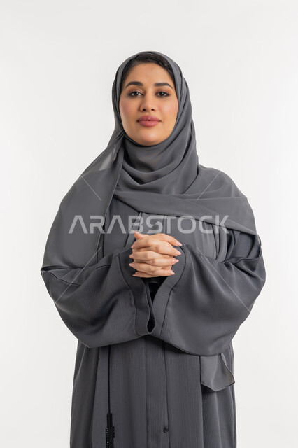 الاناقة و الاهتمام بالمظهر الخارجي، مفهوم النعومة والأنوثة, بورتريه لامرأة عربيه خليجية إماراتية محجبة مبتسمة ترتدي العباءه السوداء تقف باستقامة وأيدي متشابكة, شابة سعودية تنظر الى الكاميرا بايماءات الثقة بالنفس, خلفية بيضاء