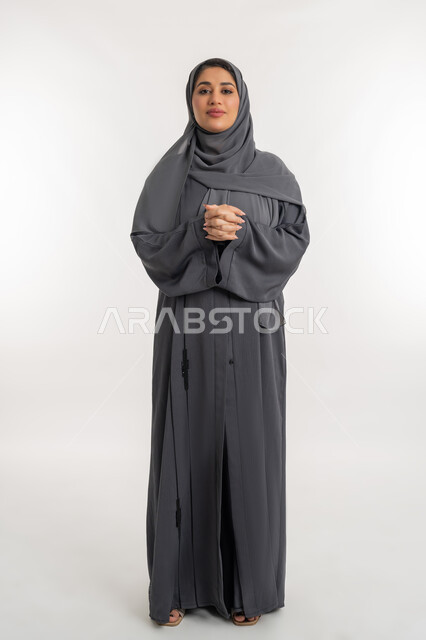 مفهوم الاناقة و الاهتمام بالمظهر الخارجي و الأنوثة, بورتريه لامرأة عربيه إماراتية محجبة مبتسمة ترتدي العباءه السوداء تقف باستقامة وأيدي متشابكة, شابة سعودية تنظر الى الكاميرا بايماءات الثقة بالنفس, الطول الكامل للجسم, خلفية بيضاء