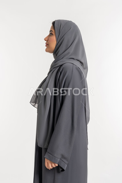Standing straight and looking to the left with gestures of self-confidence, concern for external appearance, concept of elegance and femininity, side portrait of a veiled Emirati Arab woman wearing a black abaya, a young Saudi woman looking at something, white background
