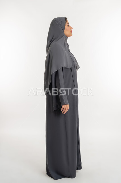Concern about appearance, concept of elegance and femininity, side portrait of a veiled Emirati Arab woman wearing a black abaya, a young Saudi woman looking at something, standing straight and looking to the right with gestures of self-confidence, full body length, white background