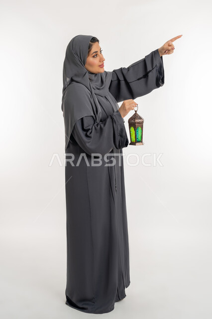 الإستعداد لقدوم شهر رمضان، المناسبات والأعياد الدينية، العبادة والتقرب إلى الله، أجواء السكينة والروحانية، بورتريه لإمراة عربية خليجية إماراتية محجبة ترتدي العباءة تحمل بيدها فانوس معدني، شابة سعودية تشير بإصبع السبابة الى شيء ما، الطول الكامل للجسم، خلفية بيضاء