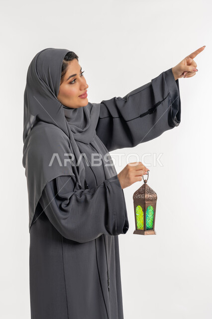 أجواء السكينة والروحانية، بورتريه لإمراة عربية خليجية إماراتية محجبة ترتدي العباءة تحمل بيدها فانوس معدني، شابة سعودية تشير بإصبع السبابة الى شيء ما، الإستعداد لقدوم شهر رمضان المبارك ، المناسبات والأعياد الدينية، العبادة والتقرب إلى الله، خلفية بيضاء