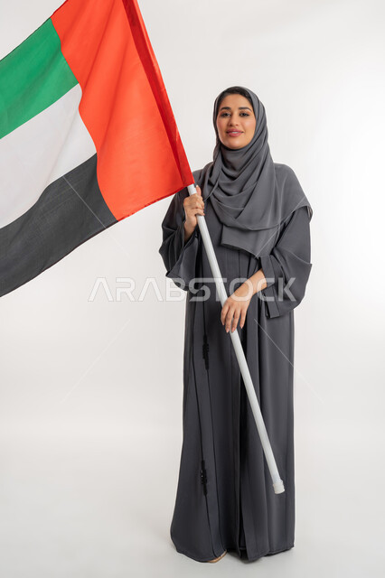 النظر الى الكاميرا بايماءات الاعتزاز والثقة، هوية اليوم الوطني الاماراتي 51، احياء ذكرى استقلال الإمارات، بورتريه لامراة عربية خليجية إماراتية ترتدي العباءة تقوم بحمل علم الإمارات، الحب والانتماء للوطن، الطول الكامل للجسم، خلفية بيضاء