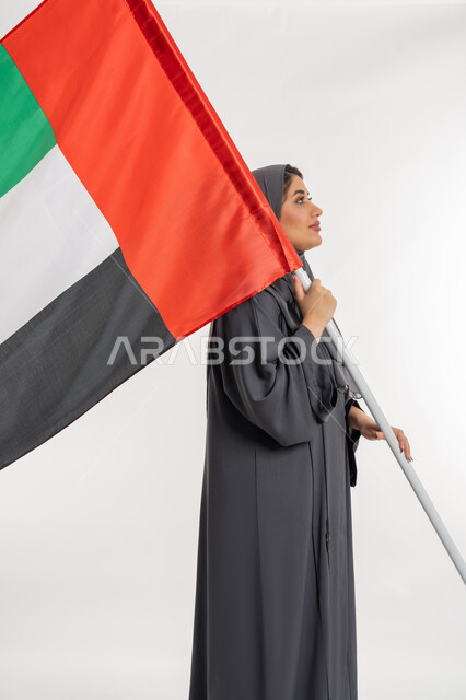 إظهار مشاعر الفخر بالهوية الاماراتية, ذكرى الإستقلال في 2 ديسمبر, حب الوطن والانتماء إليه، يوم العلم الاماراتي 3 نوفمبر, بورتريه من الجانب لامراة عربية خليجية إماراتية ترتدي العباءة تقوم بحمل علم الإمارات, الاحتفال بالعيد الوطني الاماراتي, خلفية بيضاء