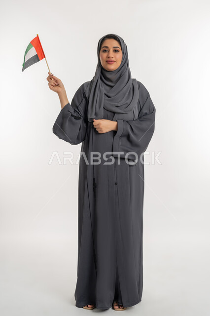 الإحتفال بالعيد الوطني الإماراتي 2 ديسمبر، إحياء ذكرى استقلال دولة الإمارات العربية المتحدة، بورتريه لامراة عربية خليجية إماراتية ترتدي عباءة سوداء تحمل علم الإمارات، الاعتزاز والفخر بإنجازات الوطن، مفهوم حب الوطن والعلم، الطول الكامل للجسم، خلفية بيضاء