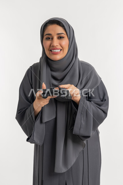 اللعب بالألعاب الترفيهية، المتعة والتسلية بالفيديو جيمز، قضاء إجازة صيفية سعيدة، استخدام التقنيات الالكترونية الحديثة, بورتريه لإمرأة عربية خليجية إماراتية محجبة ترتدي عباءة تحمل بيدها عصا التحكم، شابة سعودية بإيماءات الاستمتاع والاندماج والتفاعل, خلفية بيضاء