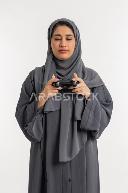 Digital entertainment world, spending free time playing video games, modern electronic technologies, portrait of a veiled Emirati Gulf Arab woman wearing an abaya holding a joystick in her hand and closing her eyes with expressions of defeat, a young Saudi woman with gestures of sadness and loss, white background
