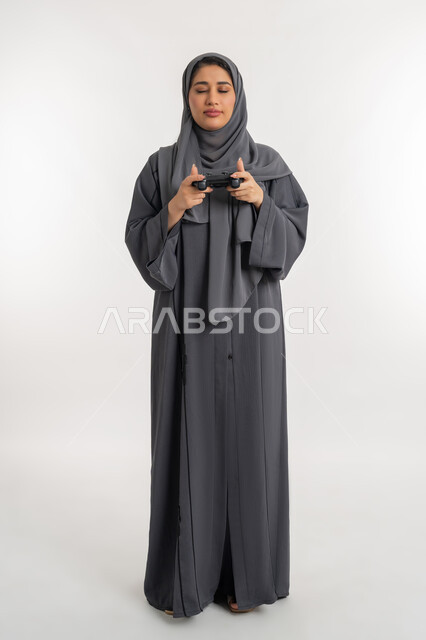 قضاء أوقات الفراغ باللعب في ألعاب الفيديو، تقنيات الكترونية حديثة, بورتريه لإمرأة عربية خليجية إماراتية محجبة ترتدي عباءة تحمل بيدها عصا التحكم وتغمض عينيها بتعابير الهزيمة، شابة سعودية بإيماءات الحزن والخسارة، الطول الكامل للجسم، خلفية بيضاء