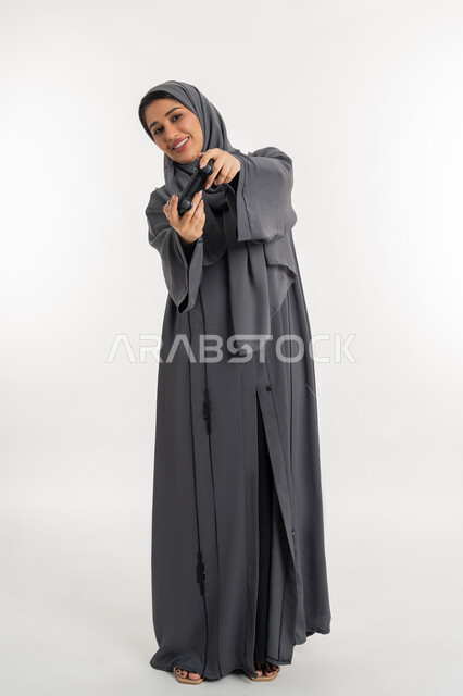 Gestures of enjoyment, integration and interaction, having a fun summer vacation, using modern electronic technologies, portrait of a veiled Emirati Gulf Arab woman wearing an abaya holding a joystick, a young Saudi woman playing with recreational games, full body length, white background