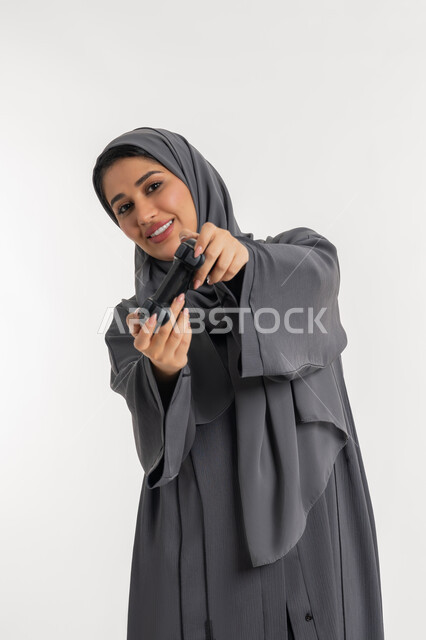 Having a happy summer vacation, using modern electronic technologies, portrait of a veiled Emirati Gulf Arab woman wearing an abaya holding a joystick, playing recreational games, a young Saudi woman enjoying video games, gestures of enjoyment, integration and interaction, white background