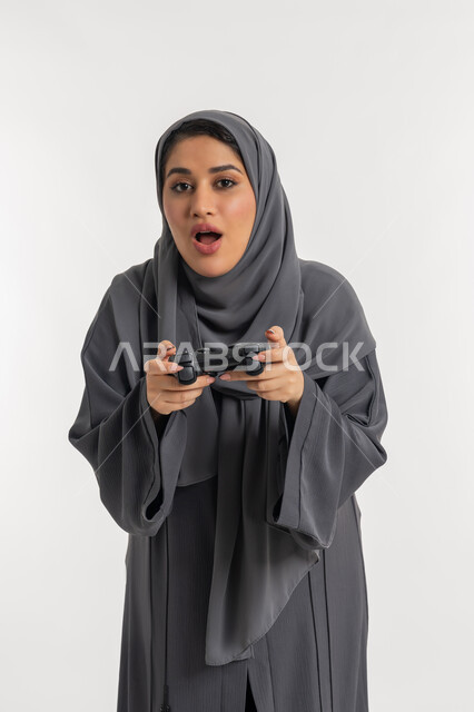 المتعة والتسلية بالفيديو جيمز، قضاء إجازة صيفية سعيدة، استخدام التقنيات الالكترونية الحديثة, بورتريه لإمرأة عربية خليجية إماراتية محجبة ترتدي عباءة تحمل بيدها عصا التحكم، شابة سعودية بإيماءات الاستمتاع والاندماج والتفاعل, اللعب بالألعاب الترفيهية، خلفية بيضاء