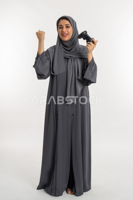 التفاعل مع الألعاب الالكترونية الترفيهية، المتعة والتسلية بالفيديو جيمز، بورتريه لإمرأة عربية خليجية إماراتية محجبة ترتدي عباءة تحمل بيدها عصا التحكم وترفع قبضتها للأعلى بتعابير الانتصار، شابة سعودية بايماءات الاندماج والتركيز، الطول الكامل للجسم، خلفية بيضاء