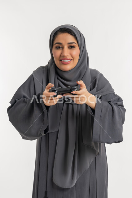 استخدام التقنيات الالكترونية الحديثة, بورتريه لإمرأة عربية خليجية إماراتية محجبة ترتدي عباءة تحمل بيدها عصا التحكم، اللعب بالألعاب الترفيهية، شابة سعودية بإيماءات الاستمتاع والاندماج والتفاعل, المتعة والتسلية بالفيديو جيمز، قضاء إجازة صيفية سعيدة، خلفية بيضاء