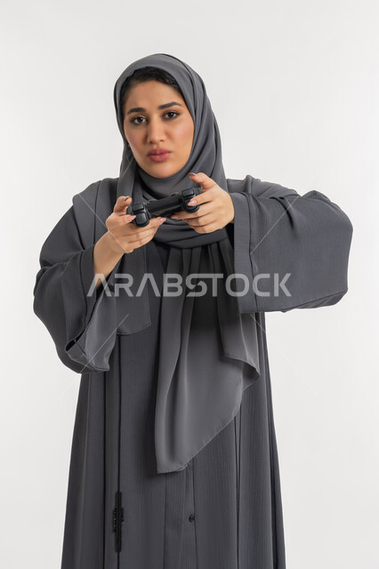 العالم الرقمي الترفيهي، قضاء أوقات الفراغ باللعب في ألعاب الفيديو الترفيهية، تقنيات الكترونية حديثة, بورتريه لإمرأة عربية خليجية إماراتية محجبة ترتدي عباءة تحمل بيدها عصا التحكم بتعابير الخوف، شابة سعودية بإيماءات الحزن والخسارة، خلفية بيضاء