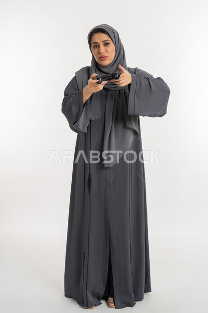 قضاء أوقات الفراغ باللعب في ألعاب الفيديو الترفيهية، تقنيات الكترونية حديثة, بورتريه لإمرأة عربية خليجية إماراتية محجبة ترتدي عباءة تحمل بيدها عصا التحكم بتعابير الخوف، شابة سعودية بإيماءات الحزن والخسارة،  العالم الرقمي الترفيهي، الطول الكامل للجسم، خلفية بيضاء