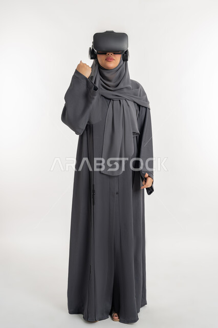 Concept of using modern technology, integration and simulation of the imaginary world, portrait of a veiled Gulf Arab Emirati woman wearing an abaya and virtual reality glasses, a young Saudi woman clenching her fist tightly with expressions of enthusiasm, victory, strength and encouragement, full body length, white background