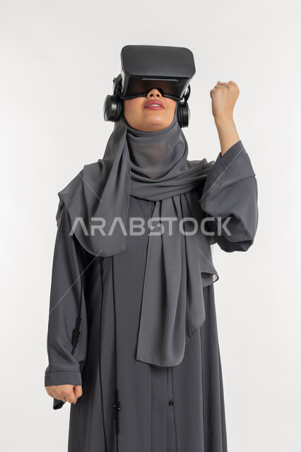 تعابير الحماس والسعادة، مفهوم استخدام التكنلوجيا والتقنيات الحديثة، الاندماج ومحاكاة العالم الخيالي, بورتريه لإمرأة عربية خليجية إماراتية محجبة ترتدي العباءة وتضع نظارة الواقع الافتراضي تغلق قبضة يدها بشدة، شابة سعودية بايماءات القوة والتشجيع، خلفية بيضاء