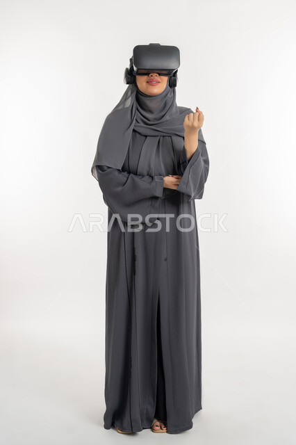 مفهوم استخدام التكنلوجيا والتقنيات الحديثة، الاندماج مع العالم الخيالي، بورتريه لإمرأة عربية خليجية إماراتية محجبة ترتدي العباءة وتضع نظارة الواقع الافتراضي تغلق قبضة يدها بشدة بتعابير الحماس، شابة سعوية بايماءات القوة والتشجيع، الطول الكامل للجسم، خلفية بيضاء