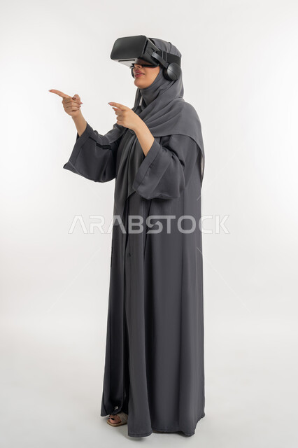 رفع اصبع السبابة والاشارة الى مكان معين، المشاهدة عبر نظارة الواقع الافتراضي, بورتريه لإمرأة عربية خليجية إماراتية محجبة ترتدي العباءة تضع نظارات ثلاثية الأبعاد, شابة سعودية تحاول النقر على شيء ما, الاندماج ومحاكاة العالم الخيالي, الطول الكامل للجسم, خلفية بيضاء